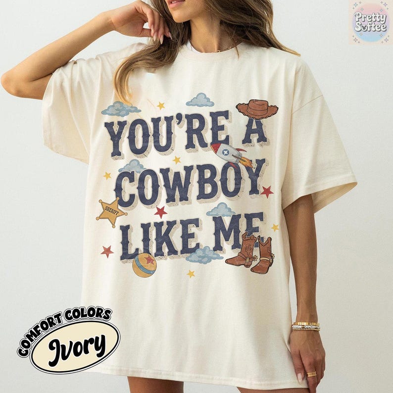 May include: An ivory t-shirt featuring the dark blue text "YOU'RE A COWBOY LIKE ME" surrounded by graphic illustrations of a brown cowboy hat, a rocket ship, a yellow sheriff star, a striped ball, and brown cowboy boots with small red stars.