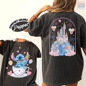 May include: Dark gray t-shirt featuring a cartoon illustration of Stitch and a castle. The front shows Stitch in a teacup with hearts. The back displays a castle scene with fireworks and balloons. The shirt has the text "Comfort Colors Pepper".