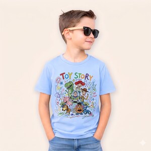 May include: A light blue t-shirt featuring the words "TOY STORY" in colorful lettering, along with cartoon character illustrations. The shirt has a crew neck and short sleeves.