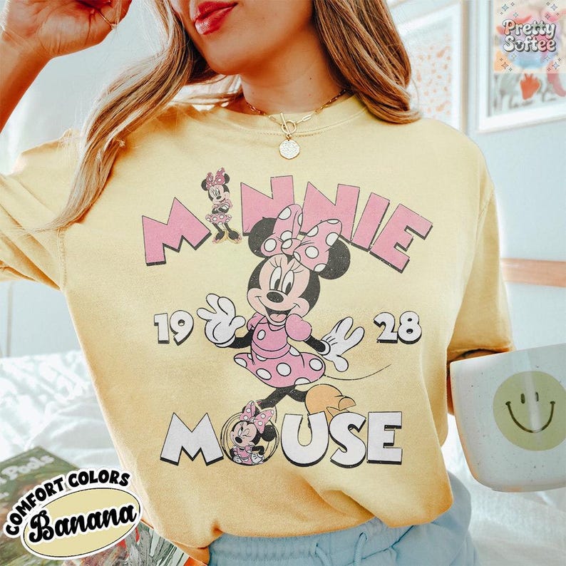 Puede incluir: Camiseta amarilla clara con Minnie Mouse con vestido y lazo de lunares rosas. El texto "MINNIE MOUSE 1928" es rosa. Las palabras "COMFORT COLORS" y "Banana" tambi&eacute;n est&aacute;n impresas en la camiseta.
