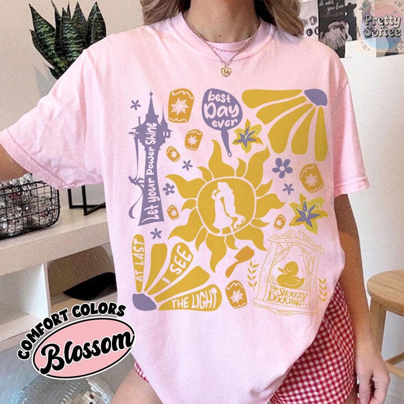 K&ouml;nnte beinhalten: Ein hellrosa T-Shirt mit einem grafischen Design mit einer Sonne, Blumen und Text. Der Text enth&auml;lt "Let your Power Shine", "Best Day Ever" und "I See The Light". Das Shirt ist mit "Comfort Colors Blossom" gekennzeichnet.