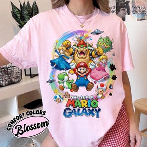 K&ouml;nnte beinhalten: Hellrosa T-Shirt mit einem farbenfrohen Grafikdesign von Super Mario Galaxy-Charakteren. Das Design zeigt Mario, Bowser und Prinzessin Peach. Der Text auf dem Shirt lautet "The Super Mario Galaxy" und "Comfort Colors Blossom".