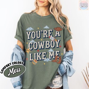 May include: A moss green short-sleeve t-shirt featuring the text "YOU'RE A COWBOY LIKE ME" in a dark blue font. The graphic includes illustrations of a brown cowboy hat, a rocket ship, a sheriff star, a ball, and brown cowboy boots.