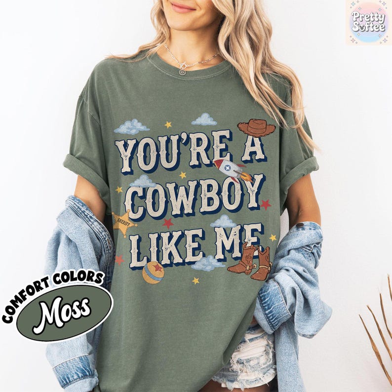 May include: A moss green short-sleeve t-shirt featuring the text "YOU'RE A COWBOY LIKE ME" in a dark blue font. The graphic includes illustrations of a brown cowboy hat, a rocket ship, a sheriff star, a ball, and brown cowboy boots.