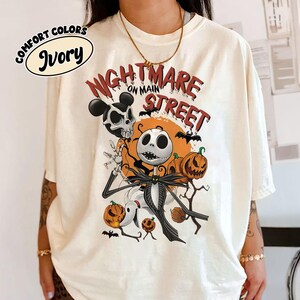 May include: Ivory-colored t-shirt featuring a graphic design with the text "Nightmare on Main Street." The design includes Jack Skellington, pumpkins, bats, and other Halloween-themed elements. The shirt is labeled "Comfort Colors."