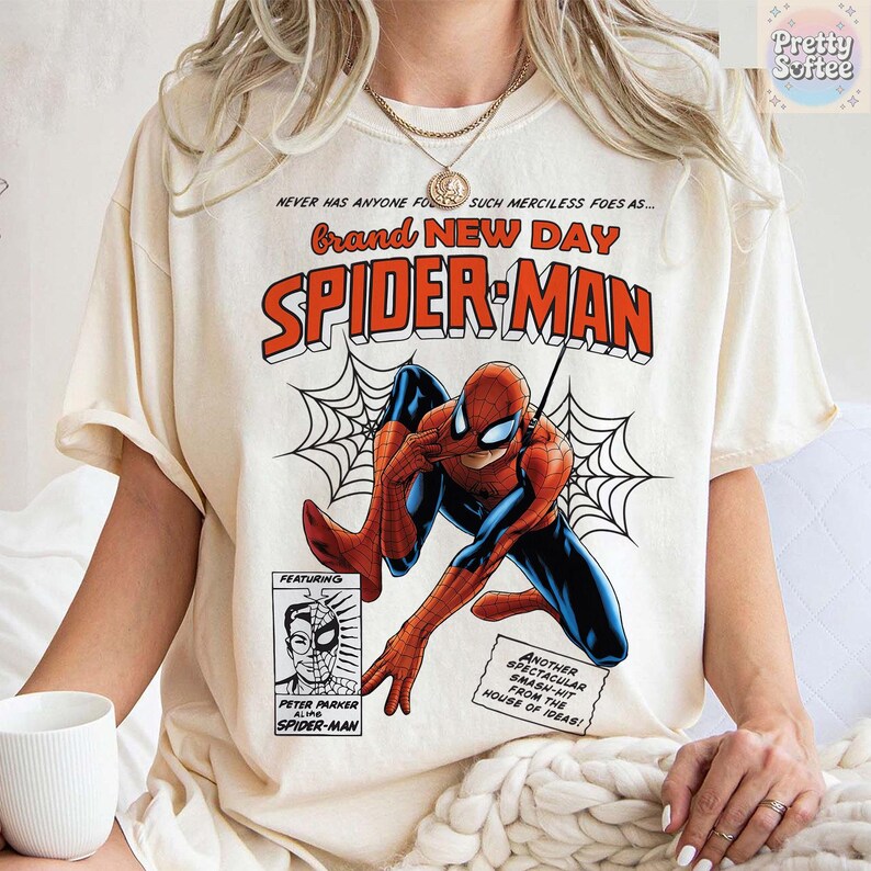 K&ouml;nnte beinhalten: Cremefarbenes T-Shirt mit einer Comic-Grafik von Spider-Man. Das Design enth&auml;lt die W&ouml;rter "Brand New Day" und "Spider-Man" in fetter, roter und orangefarbener Schrift, zusammen mit einer Zeichnung von Spider-Man in Aktion. Das Shirt hat auch den Text "Peter Parker" und "Spider-Man".