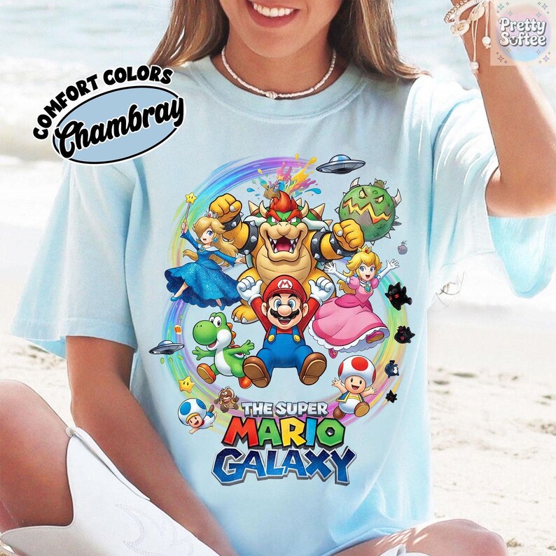 K&ouml;nnte beinhalten: Hellblaues T-Shirt mit einer farbenfrohen Grafik mit Charakteren aus dem Videospiel Super Mario Galaxy. Das Design umfasst Mario, Bowser, Prinzessin Peach, Yoshi und andere Charaktere. Der Text "THE SUPER MARIO GALAXY" wird in einer lebendigen Schriftart angezeigt.
