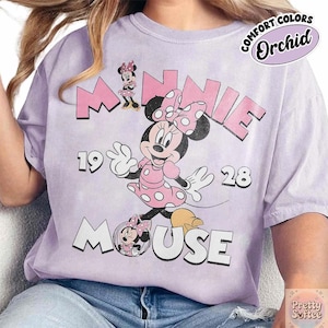 Puede incluir: Camiseta color orqu&iacute;dea con Minnie Mouse con un vestido rosa de lunares. El dise&ntilde;o incluye el texto "MINNIE MOUSE 1928" en rosa y blanco, con una est&eacute;tica vintage. La camiseta est&aacute; etiquetada como "COMFORT COLORS".