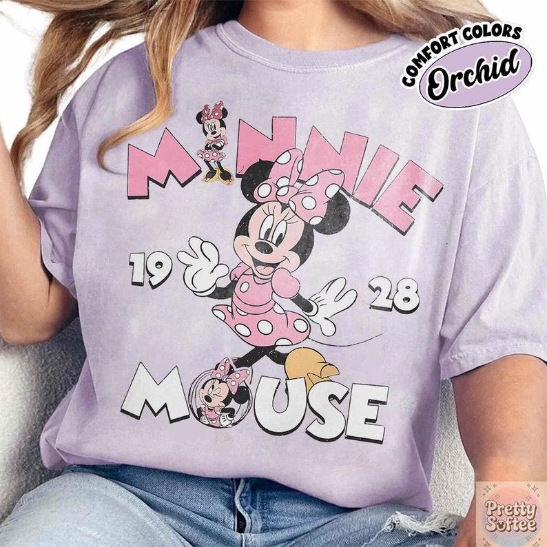 Puede incluir: Camiseta color orqu&iacute;dea con Minnie Mouse con un vestido rosa de lunares. El dise&ntilde;o incluye el texto "MINNIE MOUSE 1928" en rosa y blanco, con una est&eacute;tica vintage. La camiseta est&aacute; etiquetada como "COMFORT COLORS".