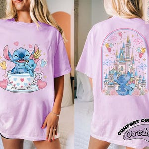 May include: A light purple t-shirt featuring a cartoon Stitch character in a teacup with hearts and balloons. The back of the shirt shows Stitch in front of a castle with fireworks. The shirt has the words "Pretty Softee" and "Comfort Colors Orchid".
