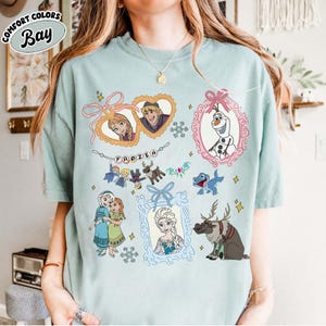 May include: A light blue t-shirt featuring various characters from the animated film "Frozen." The design includes framed portraits of Anna, Kristoff, Elsa, and Olaf, along with other characters and the word "FROZEN."