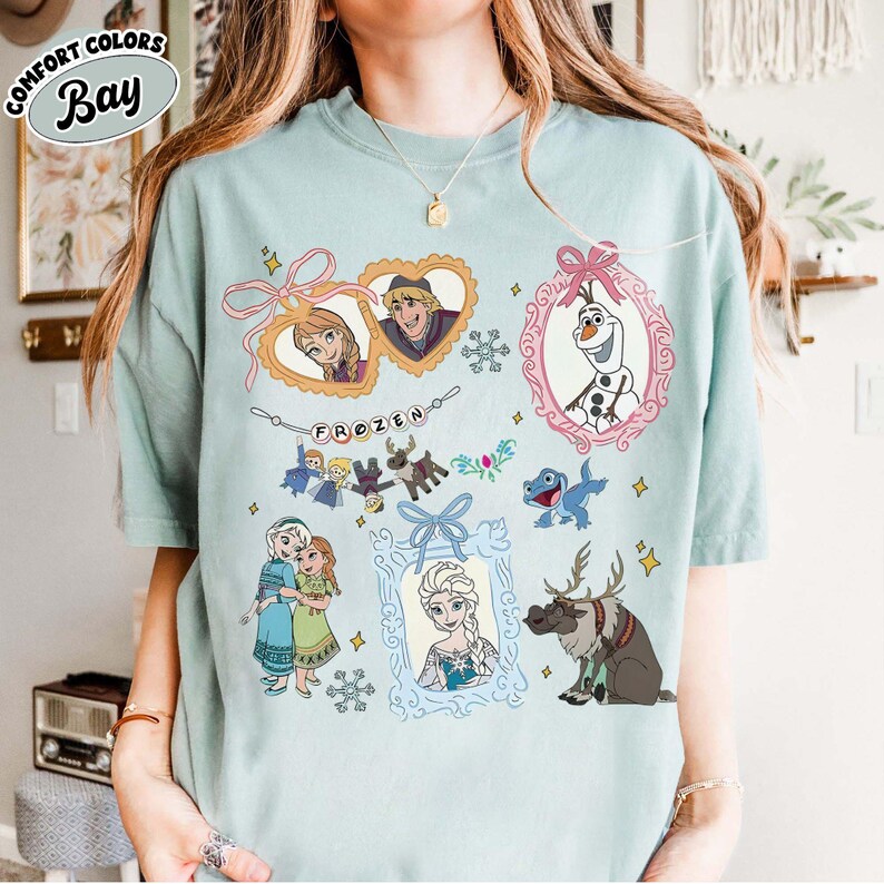 May include: A light blue t-shirt featuring various characters from the animated film "Frozen." The design includes framed portraits of Anna, Kristoff, Elsa, and Olaf, along with other characters and the word "FROZEN."