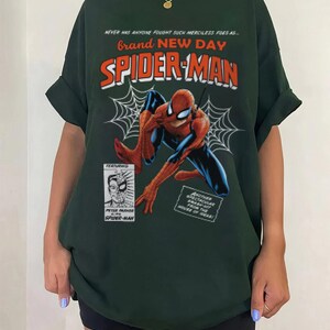 K&ouml;nnte beinhalten: Forstgr&uuml;nes T-Shirt mit einem Spider-Man-Motiv. Das Design enth&auml;lt die W&ouml;rter "Brand New Day" und "Spider-Man" in Rot und Orange, mit einer Comic-Illustration von Spider-Man und einem Spinnennetz.