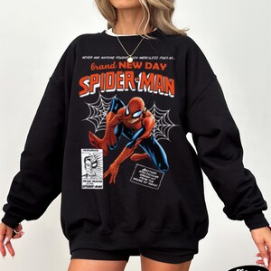 K&ouml;nnte beinhalten: Schwarzes Sweatshirt mit einem Spider-Man-Comic-Design. Die Grafik zeigt die Worte "Brand New Day" und "Spider-Man" in Rot und Orange, zusammen mit einer Illustration von Spider-Man. Das Sweatshirt hat einen wei&szlig;en Kragen.