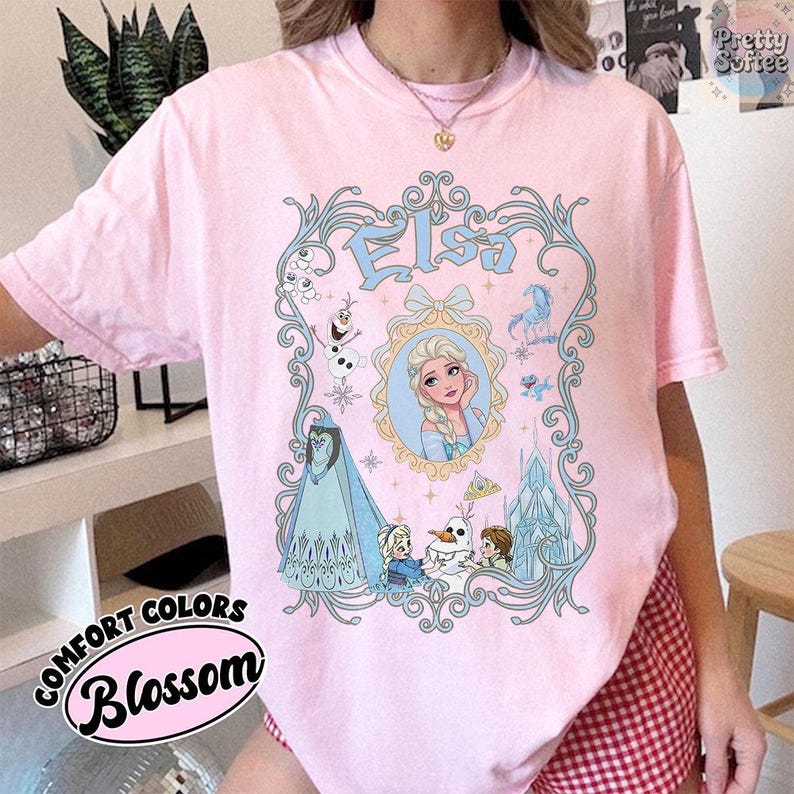 Pu&ograve; includere: T-shirt rosa chiaro con un'illustrazione dettagliata di Elsa di Frozen, con il nome "Elsa" in blu. Il design include immagini di Olaf, un castello e altri personaggi. Le parole "Comfort Colors" e "Blossom" sono stampate.