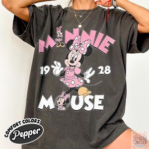 Puede incluir: Camiseta gris oscuro con un gr&aacute;fico de Minnie Mouse rosa y el texto "MINNIE MOUSE 1928". La camiseta tambi&eacute;n tiene el texto "COMFORT COLORS Pepper" en un &oacute;valo blanco.