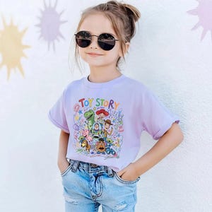 May include: A young girl wearing a light purple t-shirt with the words "TOY STORY" in colorful letters. The shirt features a graphic of characters from the animated movie. She is also wearing blue jeans and black sunglasses.