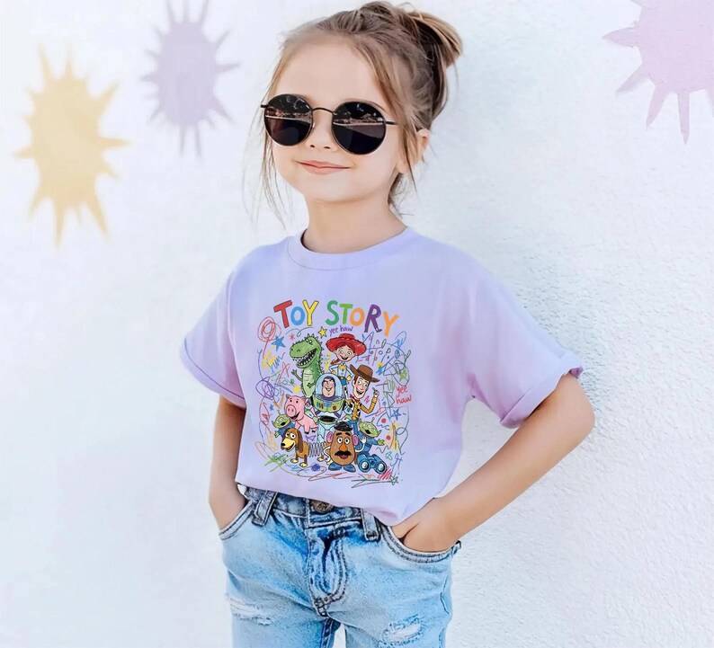 May include: A young girl wearing a light purple t-shirt with the words "TOY STORY" in colorful letters. The shirt features a graphic of characters from the animated movie. She is also wearing blue jeans and black sunglasses.