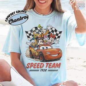 K&ouml;nnte beinhalten: Hellblaues T-Shirt mit einer Grafik von Disney-Figuren in einem Rennwagen. Das Design umfasst Micky Maus, Donald Duck, Goofy sowie Minnie und Daisy Duck. Die Worte "SPEED TEAM 1928" sind darunter gedruckt.