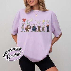 Puede incluir: Una camiseta Comfort Colors de color lila claro con un dise&ntilde;o de dibujos animados de varios personajes animales sosteniendo ramos de globos. La camiseta tiene la palabra "Orchid" impresa en una pegatina en la esquina inferior izquierda.