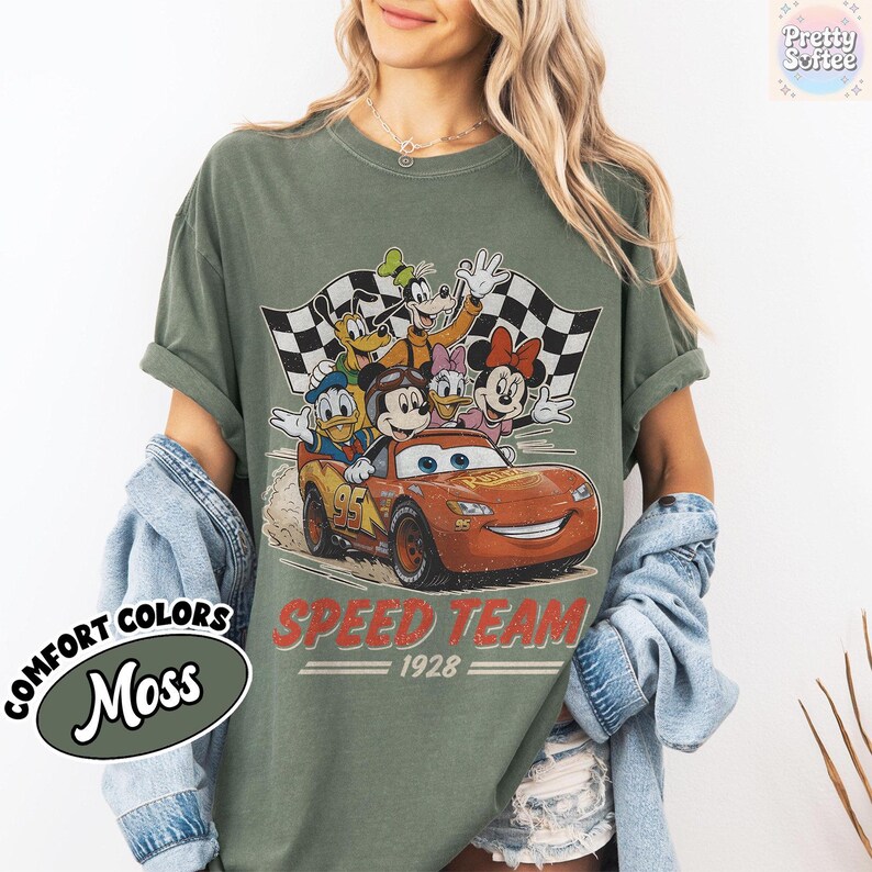 K&ouml;nnte beinhalten: Moosgr&uuml;nes T-Shirt mit einem Aufdruck von Disney-Figuren in einem Rennwagen und dem Text "SPEED TEAM 1928". Das Design umfasst Micky Maus, Donald Duck, Goofy, Daisy Duck und Minnie Maus. Das Shirt ist im Comfort-Colors-Stil.