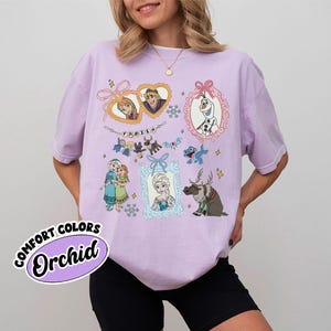 May include: A light purple t-shirt featuring various characters from the animated film "Frozen." The design includes framed portraits of Anna, Elsa, Kristoff, and Olaf, along with snowflakes and the word "Frozen." The shirt also has the words "Comfort Colors" and "Orchid" on it.