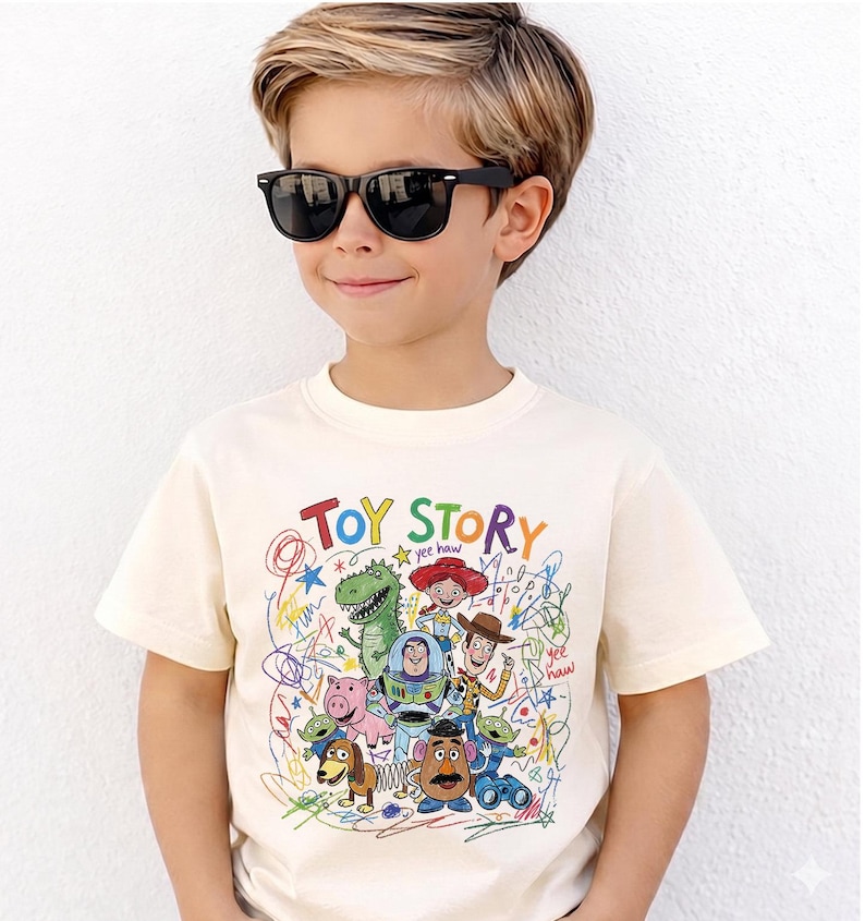 May include: Cream-colored t-shirt featuring the words "TOY STORY" in colorful letters, along with cartoon characters and scribbled designs. The shirt is worn by a young person wearing black sunglasses.