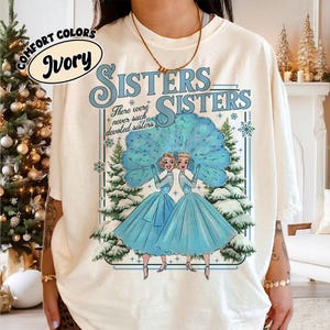 Può includere: T-shirt color avorio con le parole "Sisters Sisters" in blu. La maglietta presenta una grafica di due donne in abiti blu con una scena di foresta invernale. È incluso anche il testo "There were never such devoted sisters".