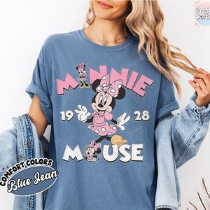 Puede incluir: Camiseta azul vaquero Comfort Colors con un gr&aacute;fico de Minnie Mouse. El dise&ntilde;o incluye la palabra "MINNIE" en letras rosas, el a&ntilde;o "1928" y la palabra "MOUSE". Minnie Mouse est&aacute; representada con un vestido rosa de lunares.