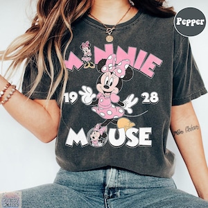 Puede incluir: Camiseta gris oscuro con Minnie Mouse con un vestido rosa con lunares blancos y un lazo a juego. La camiseta tiene el texto "MINNIE MOUSE 1928" en rosa y blanco. Una camiseta gr&aacute;fica de estilo vintage.