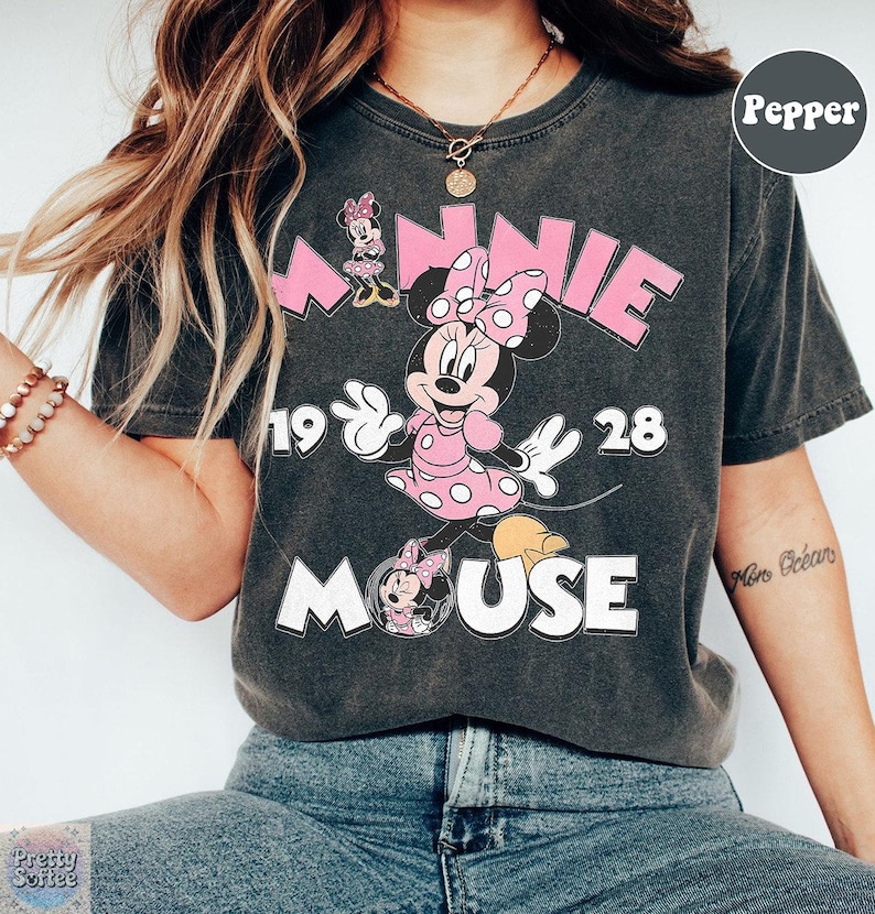 Puede incluir: Camiseta gris oscuro con Minnie Mouse con un vestido rosa con lunares blancos y un lazo a juego. La camiseta tiene el texto "MINNIE MOUSE 1928" en rosa y blanco. Una camiseta gr&aacute;fica de estilo vintage.