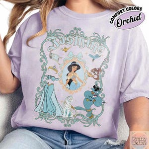 Pu&ograve; includere: Una maglietta lilla con una grafica di Jasmine. Il design include un ritratto di Jasmine, il suo abito e altri personaggi del film. La maglietta ha le parole "Jasmine" e "Comfort Colors Orchid" stampate.