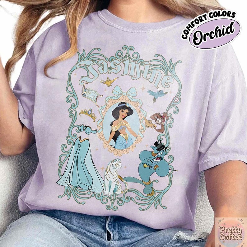 Pu&ograve; includere: Una maglietta lilla con una grafica di Jasmine. Il design include un ritratto di Jasmine, il suo abito e altri personaggi del film. La maglietta ha le parole "Jasmine" e "Comfort Colors Orchid" stampate.
