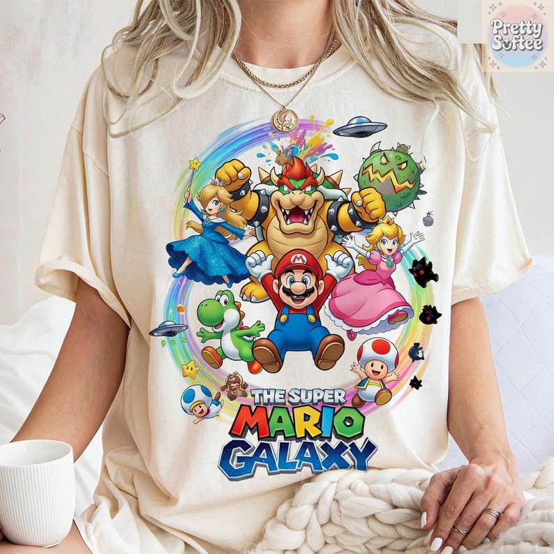 K&ouml;nnte beinhalten: Cremefarbenes T-Shirt mit einer farbenfrohen Grafik von Mario und anderen Charakteren aus dem Spiel Super Mario Galaxy. Das Design umfasst Mario, Prinzessin Peach, Yoshi, Bowser und andere Spielelemente.