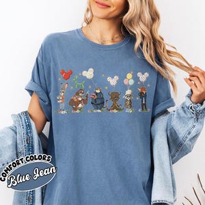 Puede incluir: Camiseta azul Comfort Colors con una ilustraci&oacute;n caprichosa de animales de dibujos animados sosteniendo globos. El dise&ntilde;o incluye un conejo, un perezoso, un oso, un toro, una cabra y un zorro. La camiseta tambi&eacute;n tiene las palabras "Comfort Colors Blue Jean".