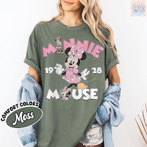 Puede incluir: Camiseta verde oliva con un gr&aacute;fico de Minnie Mouse con un vestido de lunares rosas y un lazo. La camiseta tiene el texto "MINNIE 1928 MOUSE" en rosa y blanco. Parte de la colecci&oacute;n Comfort Colors.