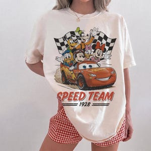 K&ouml;nnte beinhalten: Cremefarbenes T-Shirt mit einer Grafik von Disney-Figuren, darunter Micky Maus, Donald Duck und Lightning McQueen aus Cars. Das Design beinhaltet eine Zielflagge und die Worte "SPEED TEAM 1928."