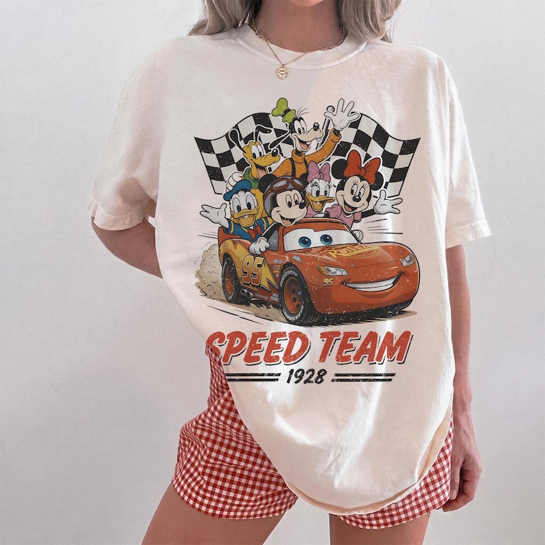 K&ouml;nnte beinhalten: Cremefarbenes T-Shirt mit einer Grafik von Disney-Figuren, darunter Micky Maus, Donald Duck und Lightning McQueen aus Cars. Das Design beinhaltet eine Zielflagge und die Worte "SPEED TEAM 1928."