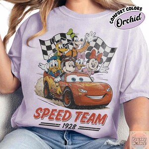 K&ouml;nnte beinhalten: Ein helllila T-Shirt mit einem Aufdruck von Disney-Figuren, darunter Micky Maus, Donald Duck und Goofy, mit einem Rennwagen und Zielflaggen. Der Text "SPEED TEAM 1928" ist darunter aufgedruckt. Das Shirt ist mit "Comfort Colors Orchid" gekennzeichnet.