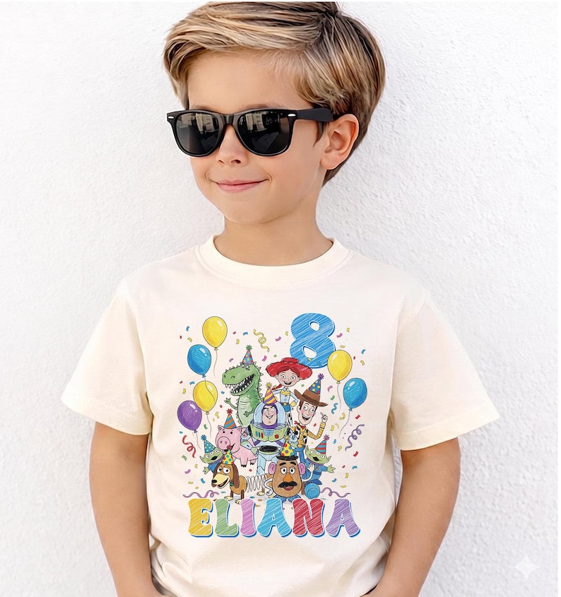 May include: Cream-colored t-shirt featuring a colorful graphic of Toy Story characters, balloons, and confetti. The number "8" and the name "ELIANA" are also displayed. The shirt is worn by a child wearing black sunglasses.