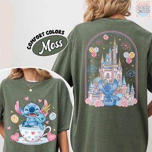 May include: Moss green t-shirt featuring a cartoon illustration of Stitch and Angel. The front shows Stitch and Angel in a teacup with hearts. The back displays a castle with fireworks and balloons. The text "COMFORT COLORS" and "Moss" are also visible.