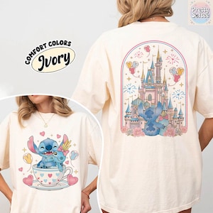 May include: Ivory-colored t-shirt featuring a cartoon illustration of Stitch and Angel in a teacup, with hearts and balloons. The back of the shirt displays a castle scene with Stitch and Angel. The text "COMFORT COLORS" and "Ivory" are also visible.