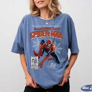 K&ouml;nnte beinhalten: Ein blaues T-Shirt mit einem Vintage-Spider-Man-Comic-Design. Die Grafik enth&auml;lt die W&ouml;rter "Brand New Day" und "Spider-Man" sowie eine Illustration des Superhelden. Das Shirt hat kurze &Auml;rmel.