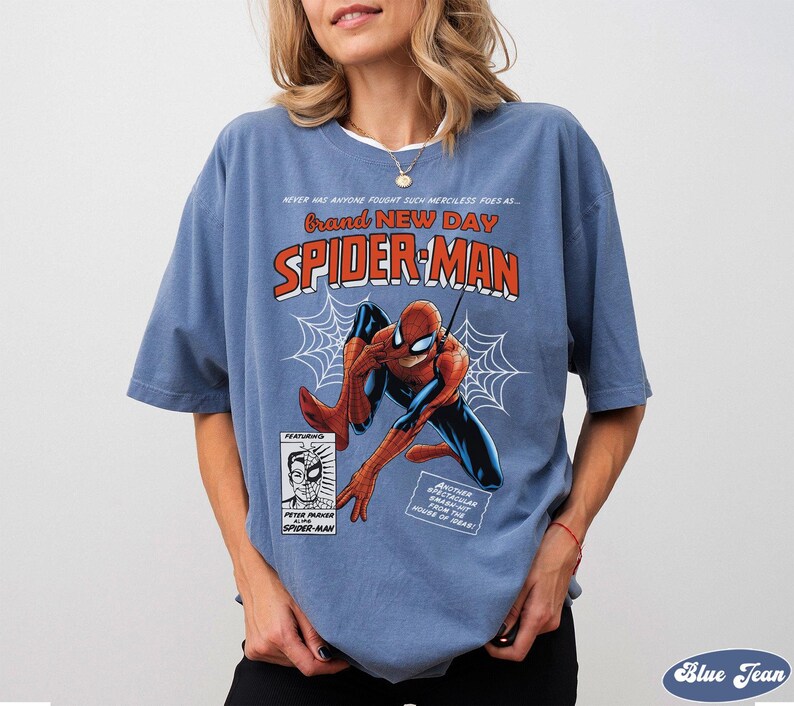 K&ouml;nnte beinhalten: Ein blaues T-Shirt mit einem Vintage-Spider-Man-Comic-Design. Die Grafik enth&auml;lt die W&ouml;rter "Brand New Day" und "Spider-Man" sowie eine Illustration des Superhelden. Das Shirt hat kurze &Auml;rmel.