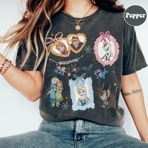 May include: Dark gray t-shirt featuring various characters and elements from the movie "Frozen." The design includes framed portraits of Anna, Elsa, Kristoff, and Olaf, along with the word "FROZEN" in a banner. Additional graphics include snowflakes and reindeer.