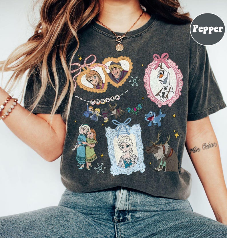 May include: Dark gray t-shirt featuring various characters and elements from the movie "Frozen." The design includes framed portraits of Anna, Elsa, Kristoff, and Olaf, along with the word "FROZEN" in a banner. Additional graphics include snowflakes and reindeer.