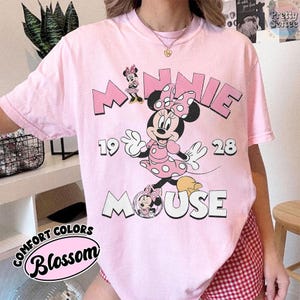 Puede incluir: Camiseta rosa claro con un dise&ntilde;o de Minnie Mouse. El dise&ntilde;o incluye la palabra "MINNIE" en rosa, los n&uacute;meros "19 28" y la palabra "MOUSE". La camiseta tambi&eacute;n tiene las palabras "COMFORT COLORS" y "Blossom" en un dise&ntilde;o circular.