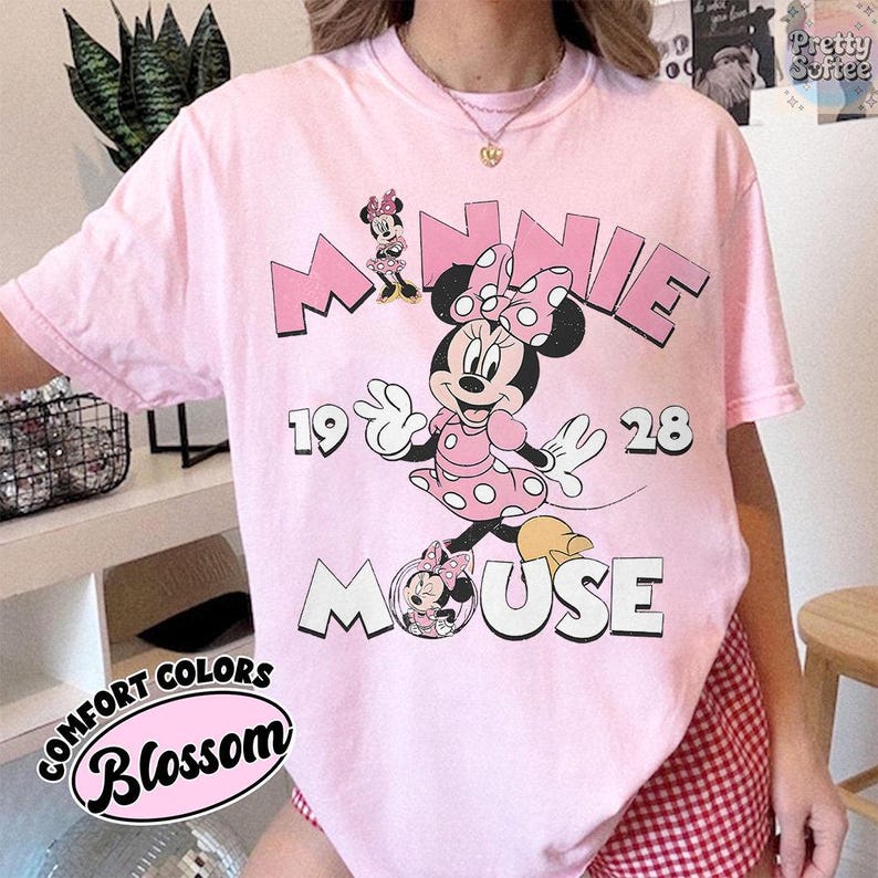 Puede incluir: Camiseta rosa claro con un dise&ntilde;o de Minnie Mouse. El dise&ntilde;o incluye la palabra "MINNIE" en rosa, los n&uacute;meros "19 28" y la palabra "MOUSE". La camiseta tambi&eacute;n tiene las palabras "COMFORT COLORS" y "Blossom" en un dise&ntilde;o circular.