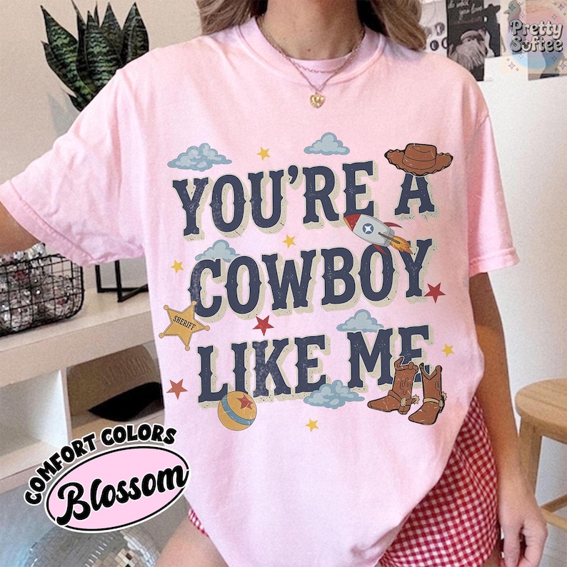 May include: A light pink t-shirt featuring the dark blue text "YOU'RE A COWBOY LIKE ME" surrounded by graphic illustrations of a brown cowboy hat, a rocket ship, a yellow sheriff star, a colorful ball, and a pair of brown cowboy boots with clouds.
