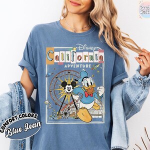 Puede incluir: Camiseta azul Comfort Colors con un dise&ntilde;o de Disney California Adventure. El dise&ntilde;o incluye a Mickey Mouse, el Pato Donald, una noria y el texto "California Adventure". Las mangas est&aacute;n dobladas.